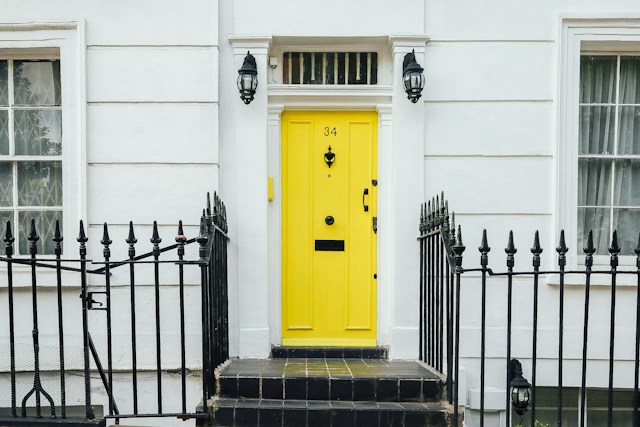 The Picture Shows the Front of a White London Terrace House with a Yellow Door and Black Railings.